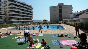 Un área de piscina con varios niveles de edificios, cielo despejado y gente tomando el sol y nadando.