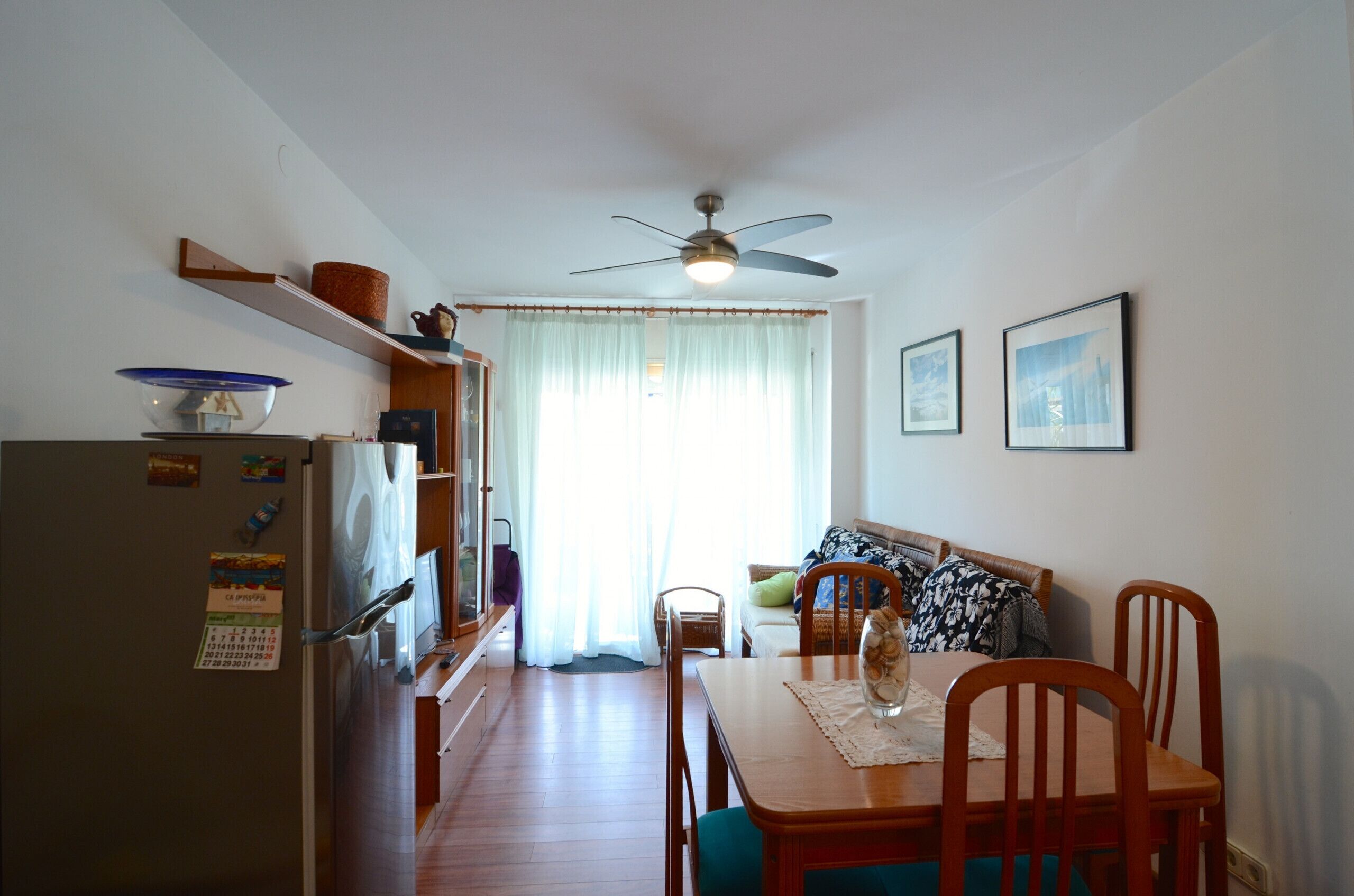 Eine kleine Küche mit Kühlschrank, einem Esstisch mit Stühlen, einem Deckenventilator und einer Couch im Hintergrund.