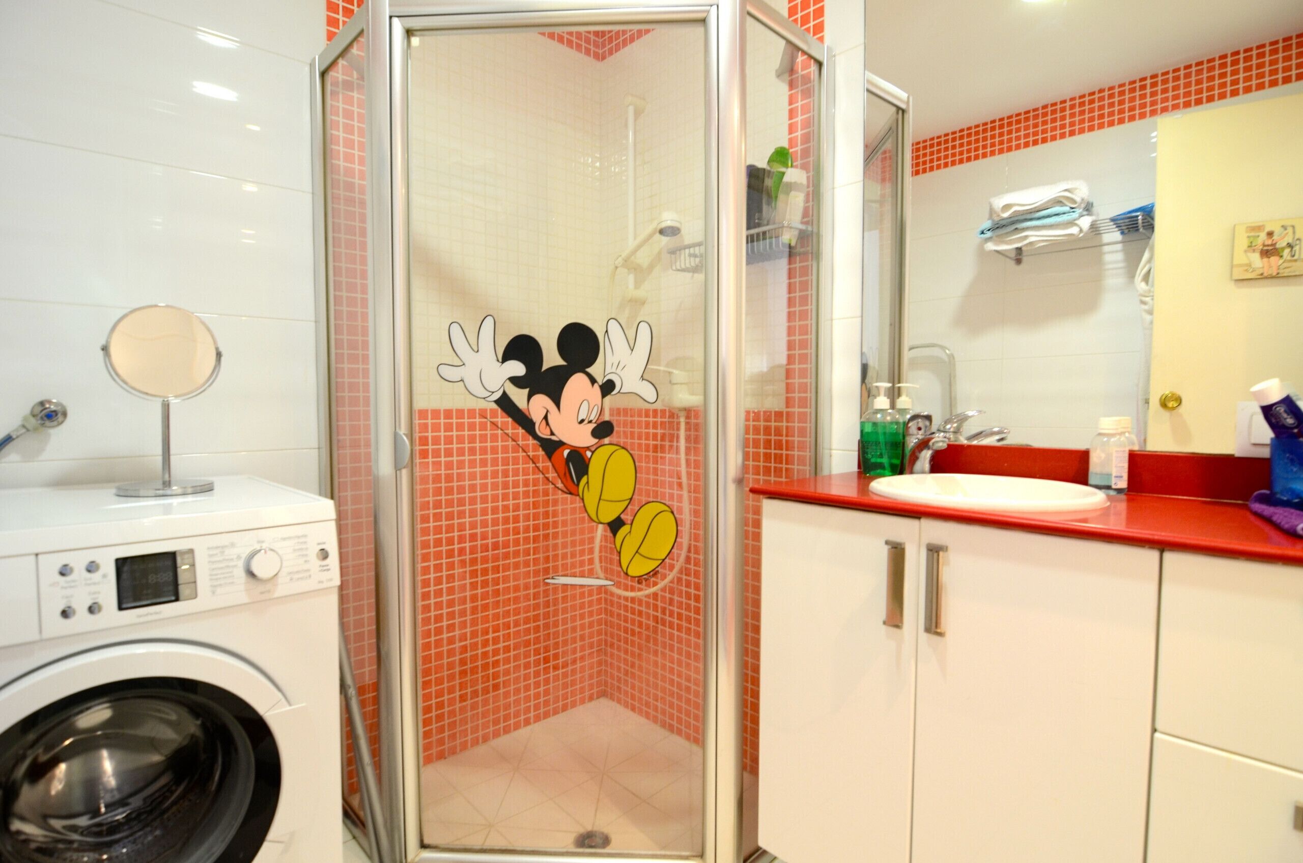 Een badkamer met een Mickey Mouse sticker op de douchedeur, een wasmachine, een wastafel met toiletartikelen en een spiegel.