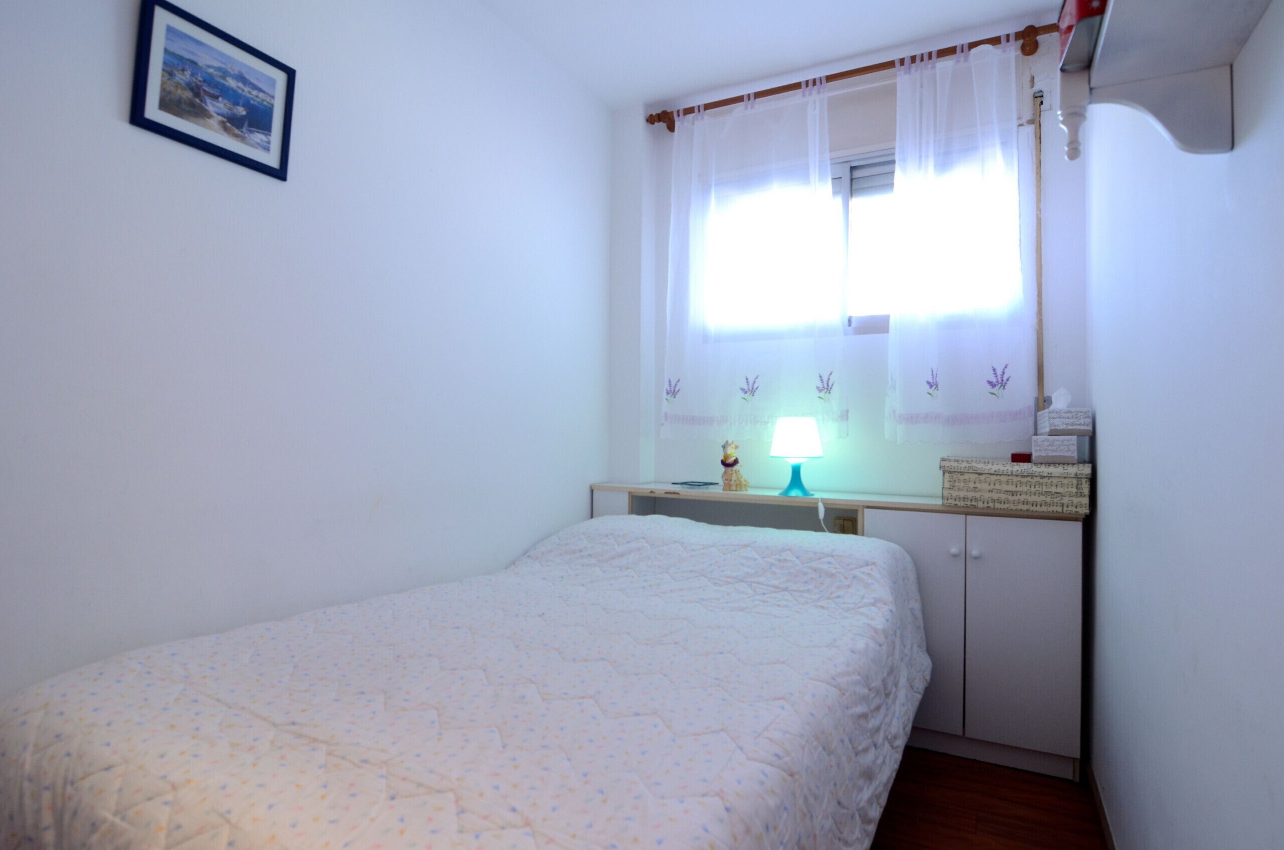 A bedroom with a bed, a nightstand, a lamp, a window with sheer curtains, and a framed picture on the wall.