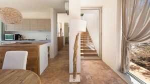 A modern kitchen with wooden cabinets, a dining area with a round table, a staircase with a white lamp, and a view through a window with curtains. - Villa Lisy by Perle di Puglia (Carovigno)