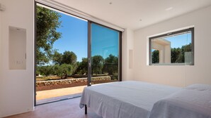 A bedroom with a large sliding glass door leading to an outdoor area, a bed with a grey bedspread, and a small window with a view of trees. - Villa Lisy by Perle di Puglia (Carovigno)