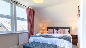 A bedroom with a bed, bedside tables, a window with curtains, a radiator, and an ottoman.