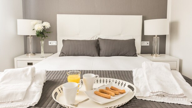 A neatly made bed with pillows, a tray with breakfast items, and a bedside table with a lamp and flowers. - Latina Suite (Madrid)