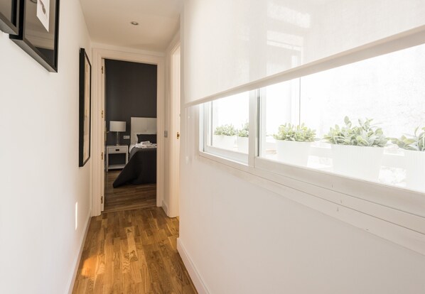 A hallway with wooden flooring, a bed, a nightstand, and a window with plants.