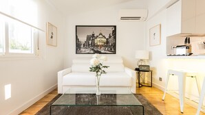 A modern living room with a glass coffee table, white sofa, and a cityscape painting. - Latina Suite (Madrid)