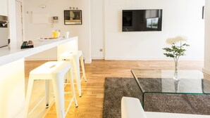 A modern kitchen with white countertops, a built-in oven, and a coffee machine. There is a dining area with a glass table and white bar stools. A flat-screen TV is mounted on the wall above the dining table. - Latina Suite (Madrid)