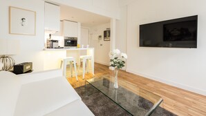 A modern living room with a glass coffee table, white sofas, a kitchen area in the background, and a mounted TV on the wall. - Latina Suite (Madrid)