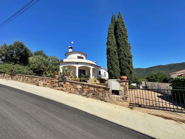 A white building with a dome roof, surrounded by stone walls and a tall cypress tree. - Villa Castell - Urb Mas Pere (Calonge)