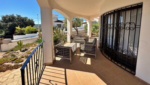 A covered balcony with a seating area, a metal gate, and a view of the street and greenery. - Villa Castell - Urb Mas Pere (Calonge)