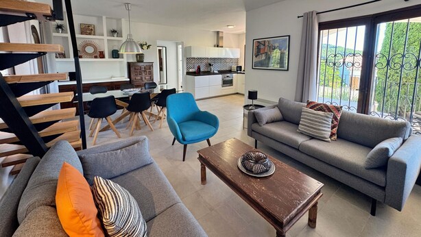 A modern living room with a grey sofa, a blue armchair, and a wooden coffee table. A staircase with wooden steps and black railings is visible on the left. - Villa Castell - Urb Mas Pere (Calonge)