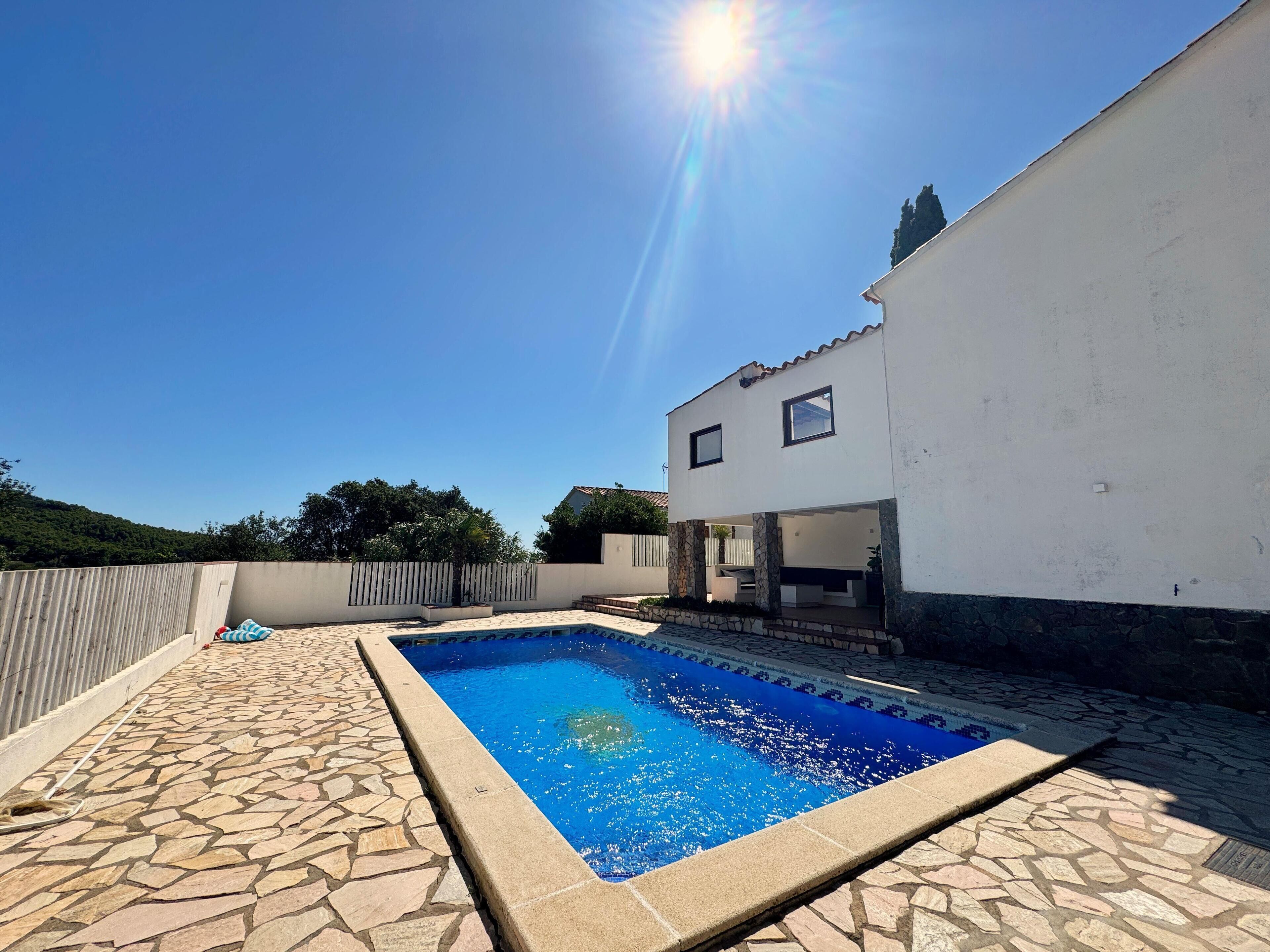 Une piscine aux bords recouverts de pierres, un bâtiment blanc doté d’un balcon et un ciel d’un bleu limpide.