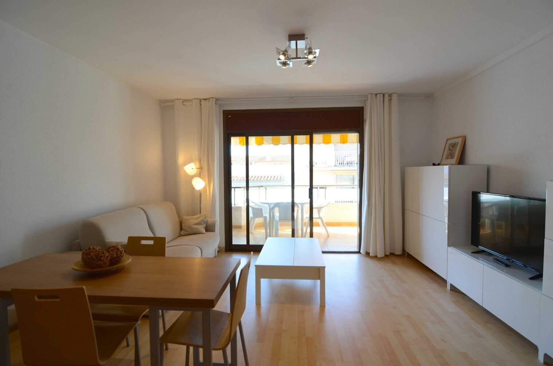 Ein modernes Wohnzimmer mit einer Couch, einem Esstisch, Stühlen, einem Fernseher und einem Balkon mit Vorhängen.