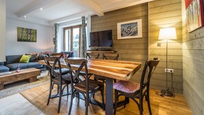 Ett mysigt vardagsrum med ett matbord i trä, bekväma sittplatser, en platt-TV och ett stort fönster med gardiner.