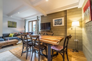 A cozy living room with a wooden dining table, comfortable seating, a flat-screen TV, and a large window with curtains.