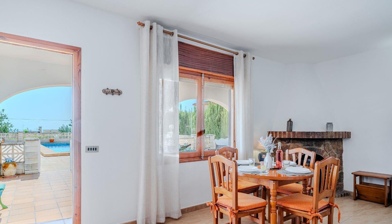 A dining area with a round table and wooden chairs, a fireplace, and a view of a swimming pool.