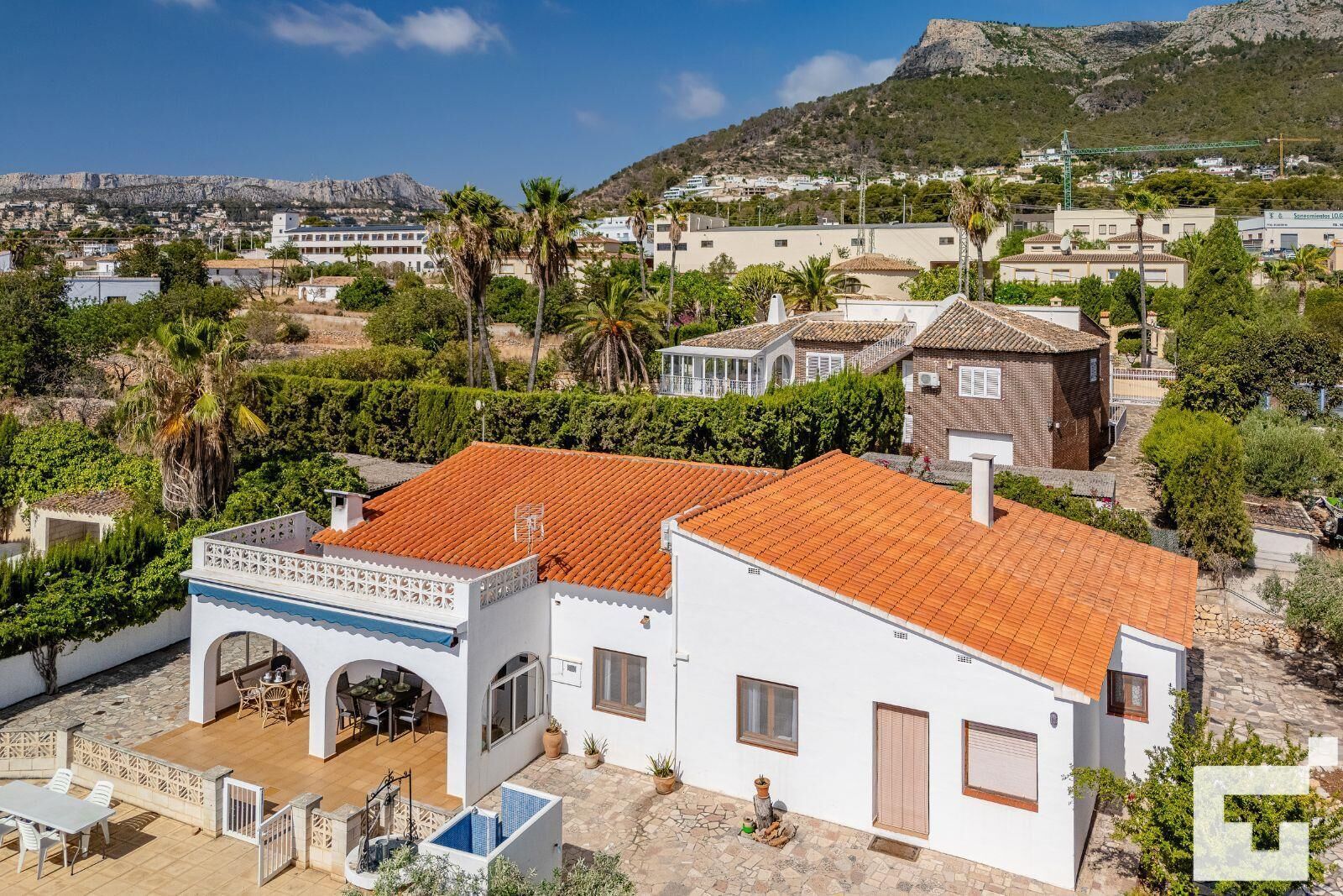 A white building with a red-tiled roof, a covered patio with outdoor furniture, and a view of a mountainous landscape in the background.