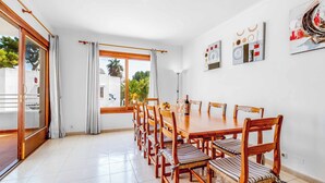 A dining area with a long wooden table and chairs, large windows with curtains, and artwork on the walls. - Villa Pou Uno - Five Bedroom Villa, Sleeps 10 (Cala d'Or)