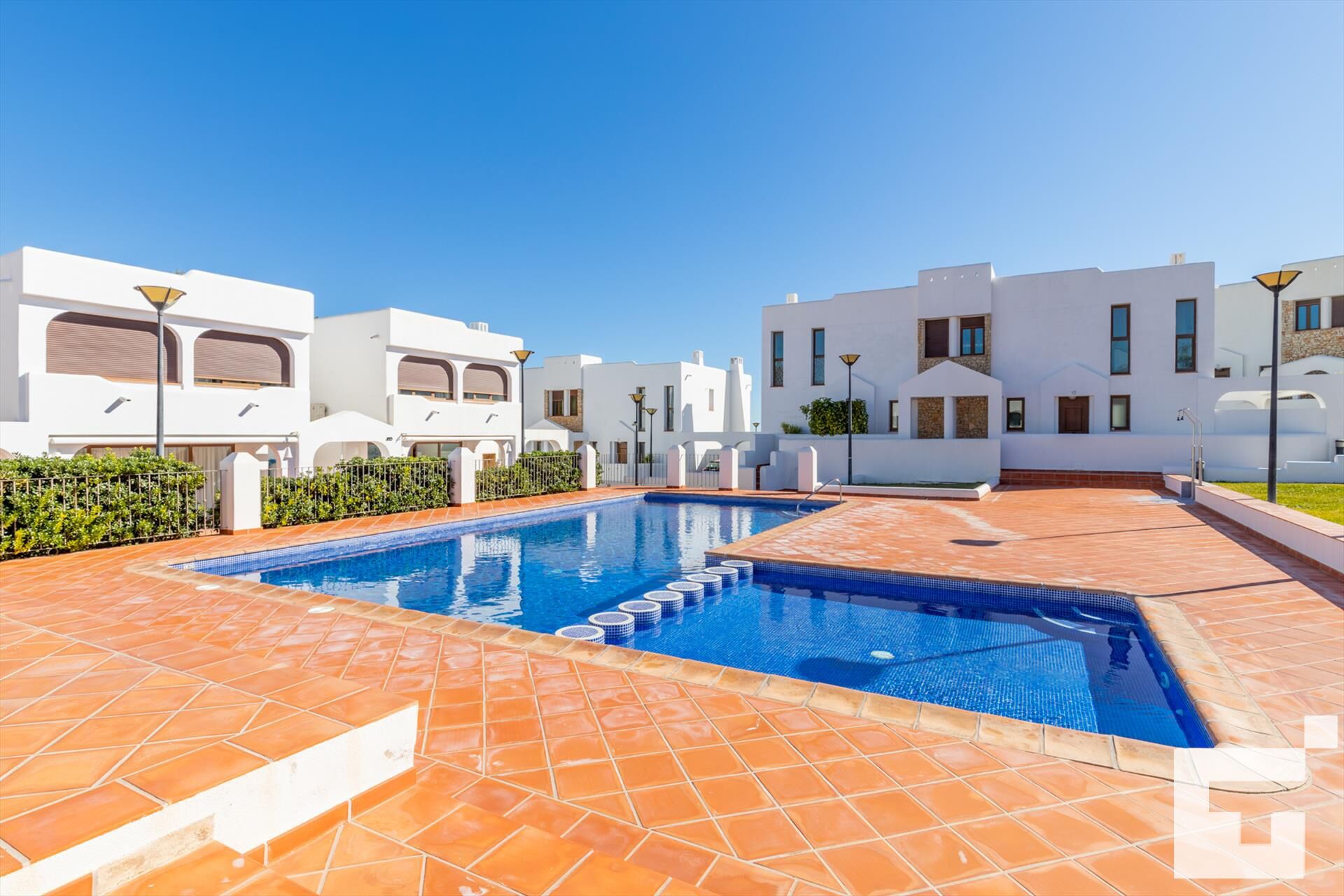 A modern pool area with a clear blue pool, surrounded by white buildings with arched windows and a tiled patio.