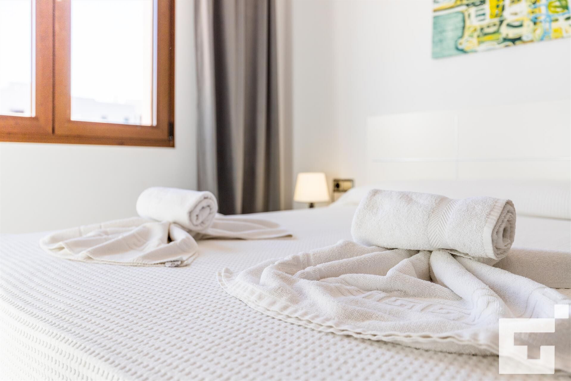 Een hotelkamer met een bed, twee opgerolde handdoeken, een lamp en een schilderij aan de muur.