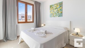 Une chambre d’hôtel avec un grand lit, une fenêtre avec des rideaux, un tableau au mur et une table de chevet avec une lampe.
