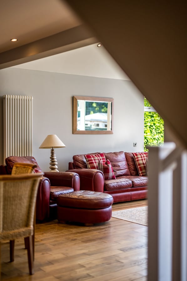 A living room with a brown leather sofa, a wooden coffee table, and a lamp. There is a window with a view of greenery outside. - Modern 2‑Bedroom St Andrews Retreat with Garden, Parking & Easy Walk to Harbour & East Sands (St. Andrews)