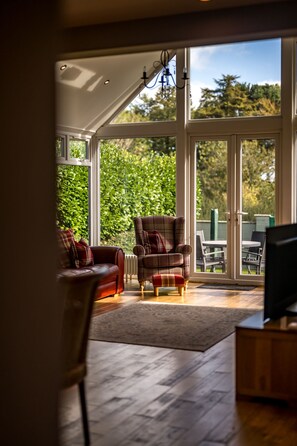 Een woonkamer met een bank, fauteuil, poef, een televisie en uitzicht op een tuin door grote glazen deuren.