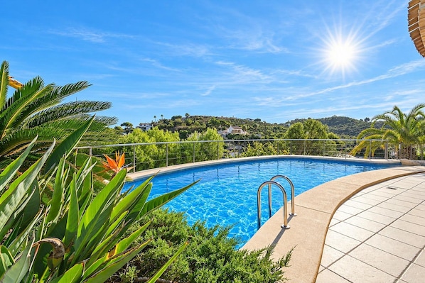 A clear blue swimming pool with a metal ladder, surrounded by tropical plants and a view of a hillside with buildings.