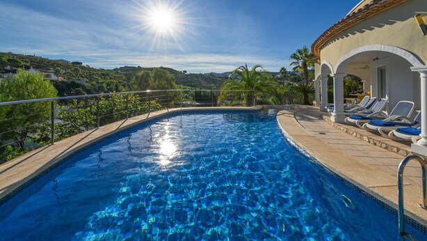A clear blue pool with a sunlit sky, surrounded by a white building with columns and a terrace with sun loungers.