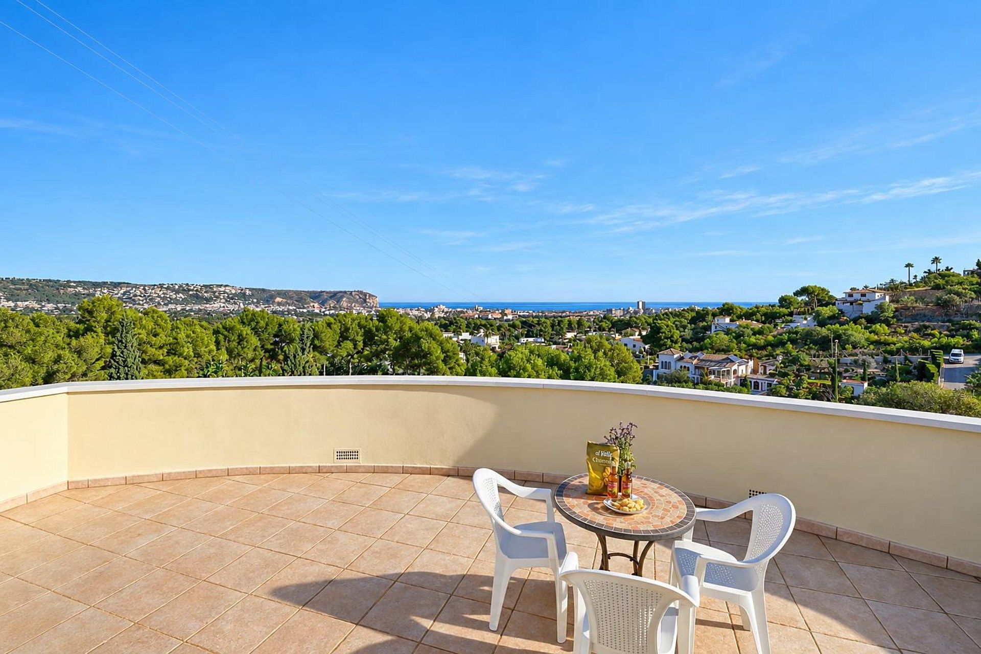Un balcon doté d’un sol carrelé, de deux chaises blanches, d’une petite table ronde ornée d’un vase et d’un paquet de chips, et offrant une vue sur une ville côtière et sur la verdure.