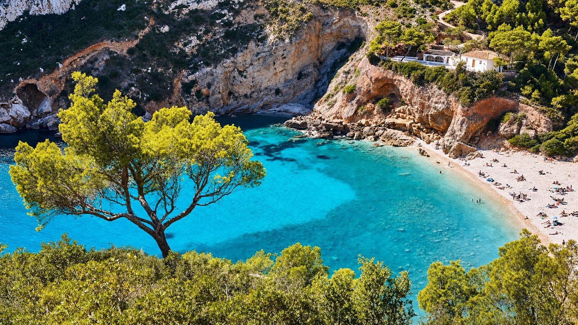 A coastal landscape with a clear blue sea, a sandy beach, and a white building with a red roof.