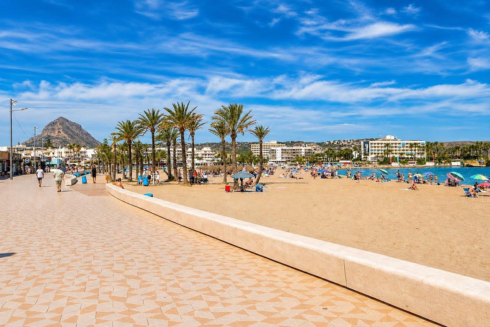 Een promenade aan het strand met palmbomen, een berg op de achtergrond en een hotel met meerdere verdiepingen.