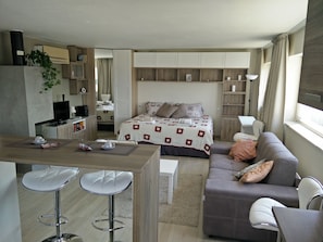 Quarto de hotel moderno e compacto, com cozinha compacta, cama com manta estampada, sofá com almofadas, pequena mesa de jantar com dois banquetes e televisão.