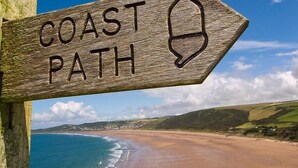 Ein Holzschild mit der Aufschrift 'COAST PATH' und einem Symbol, das eine Küstenrichtung anzeigt.