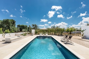 A rectangular swimming pool with clear blue water, surrounded by a tiled deck. There are two lounge chairs on one side of the pool.