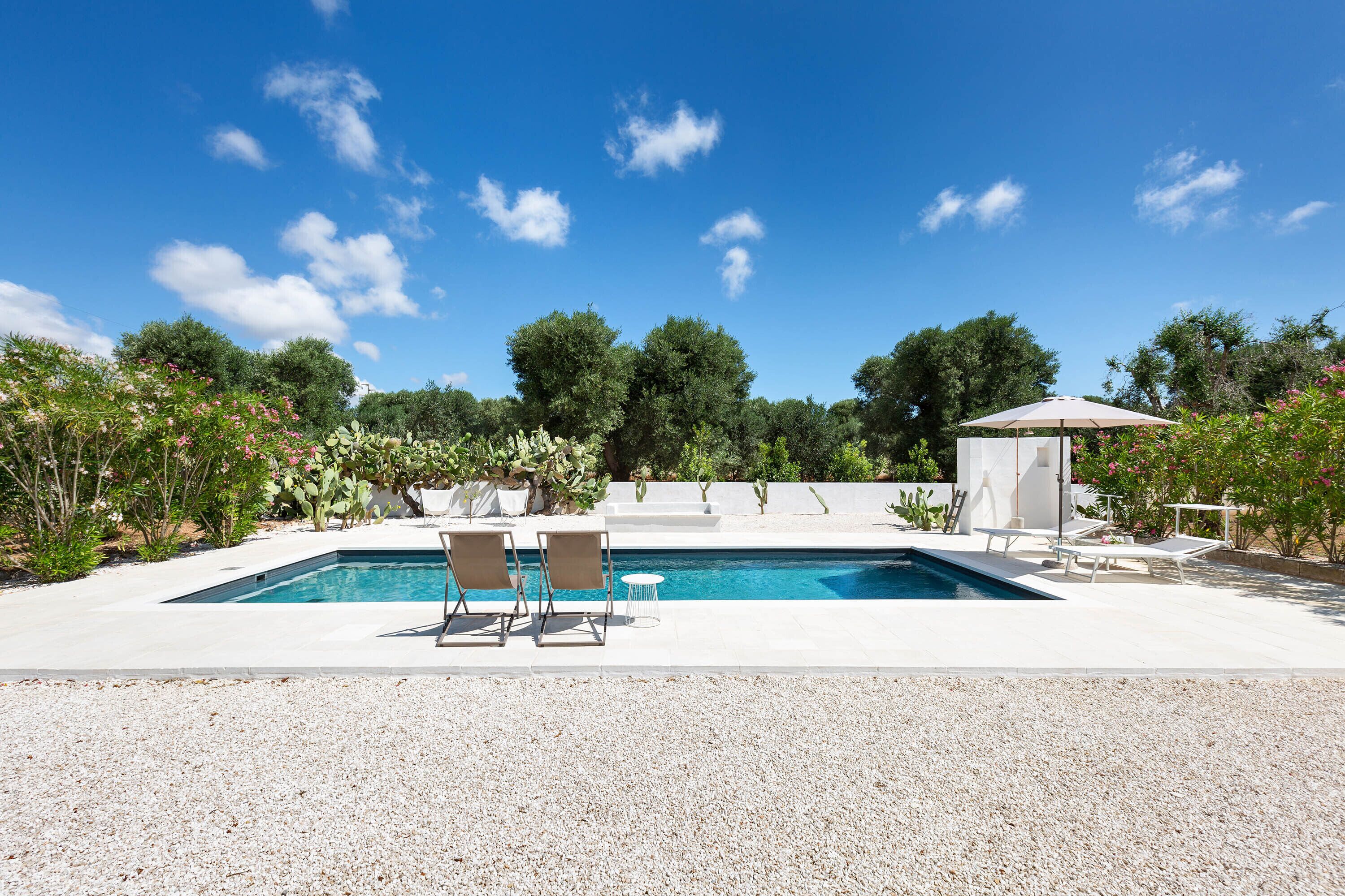 A rectangular swimming pool with two lounge chairs and a small table, surrounded by gravel and greenery.