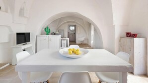 A white dining area with a table, chairs, and a plate of lemons. There is a television on a small stand to the left and a cactus decoration on the wall.