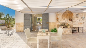 An outdoor patio with stone flooring, a table with chairs, a stone wall with a fireplace, and a hanging light fixture.