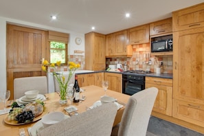 A modern kitchen with wooden cabinets, a dining table set for four, and a breakfast spread. - 2 Bed in Croyde (oc-longst) (Croyde)