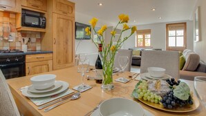 A dining area with a wooden table set for two, a vase with yellow flowers, and a fruit platter.