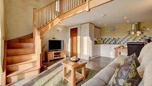A spacious living area with a wooden staircase, a flat-screen TV, a dining table, and a kitchen in the background.
