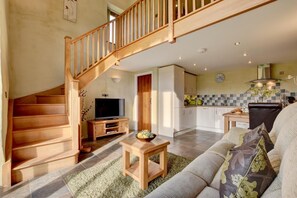 A spacious living area with a wooden staircase, a flat-screen TV, a dining table, and a kitchen in the background.
