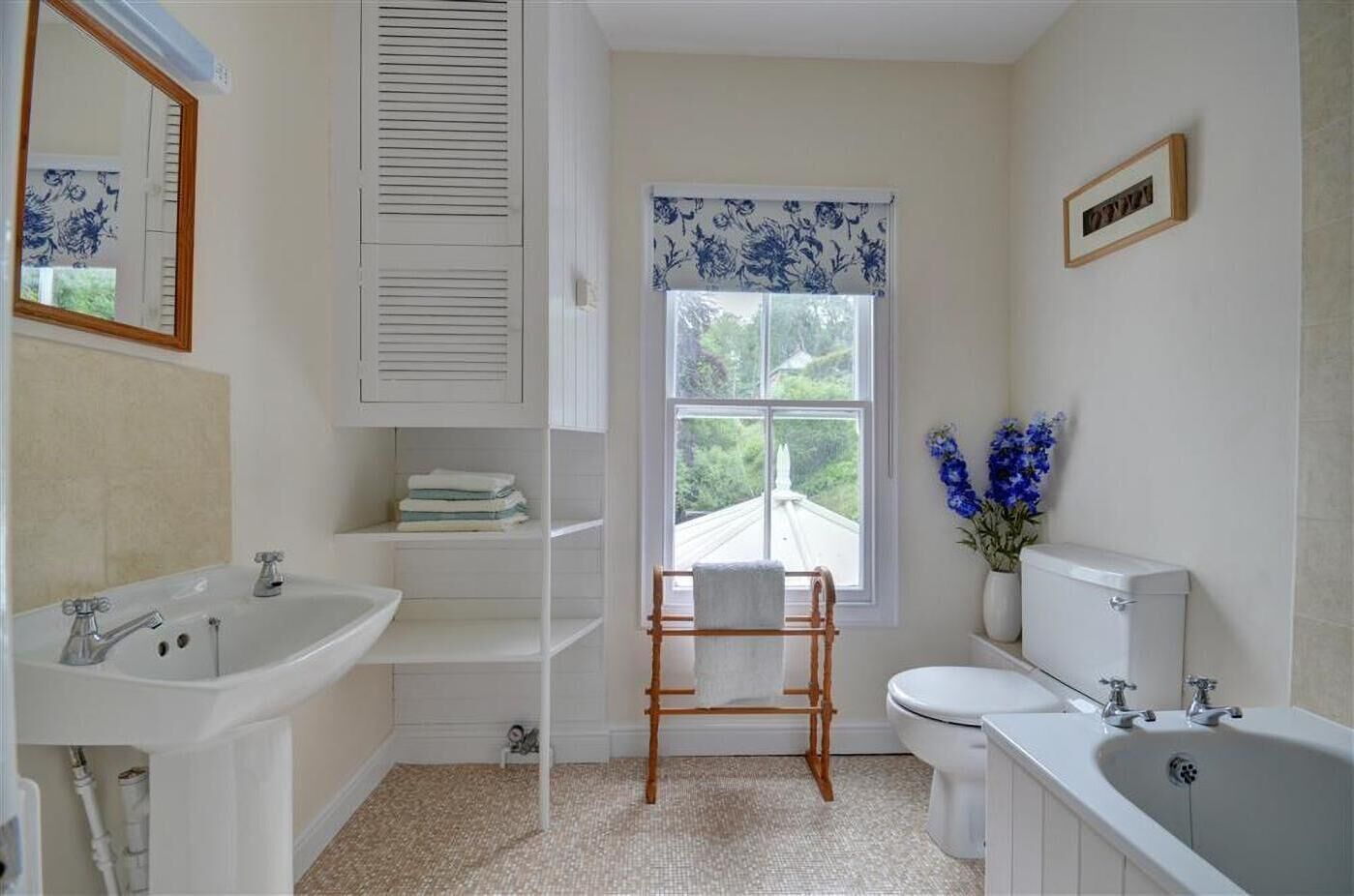 Un baño con lavamanos, inodoro y bañera blancos. Se observa una ventana con cortinas florales azules, una silla de madera y un jarrón con flores azules.