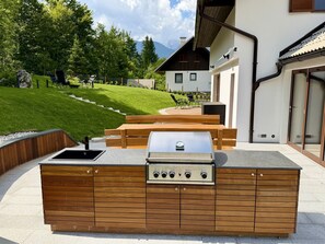 An outdoor kitchen with a built-in grill, wooden cabinetry, and a view of a house and mountains in the background.