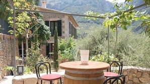 Un espace extérieur aménagé dans un style rustique, avec une table en fût de bois et des chaises en métal, adossé à un mur de pierre et offrant une vue sur un paysage montagneux.