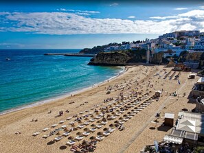 A beach with rows of sunbeds and umbrellas, a clear blue sky, and buildings in the background. - Luxury 1 Bedroom, Apartment Marina de Albufeira near Old Town (Albufeira)