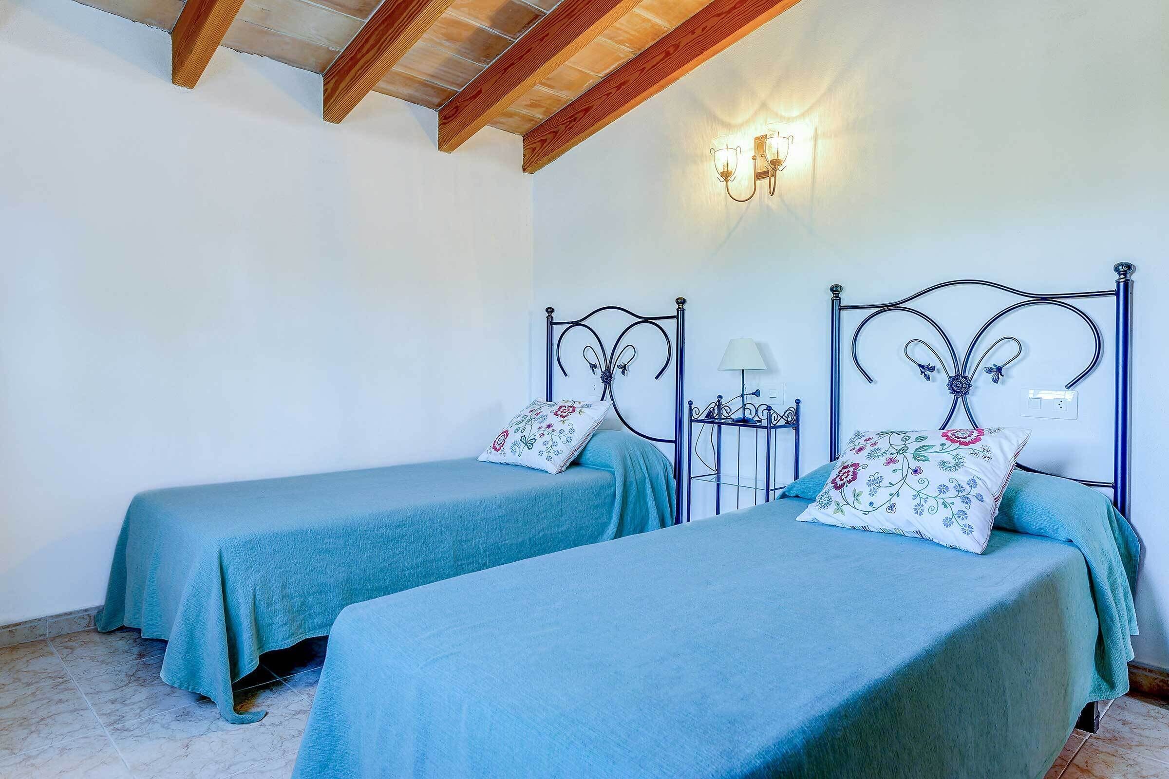 Una habitación con dos camas, cada una con un cojín floral y una colcha azul. Entre las camas hay una mesita de noche con una lámpara. El techo es de madera y hay un aplique de pared.