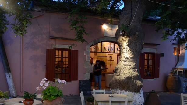 An outdoor café with stone tables and chairs, a large tree, and a building with a pink facade and wooden shutters.