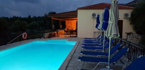 Ein Schwimmbecken mit Liegestühlen und Sonnenschirmen, eine überdachte Terrasse und ein Gebäude mit offener Eingangstür.