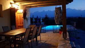 An outdoor patio with a wooden table and chairs, a pool with blue lighting, and a view of mountains in the background.